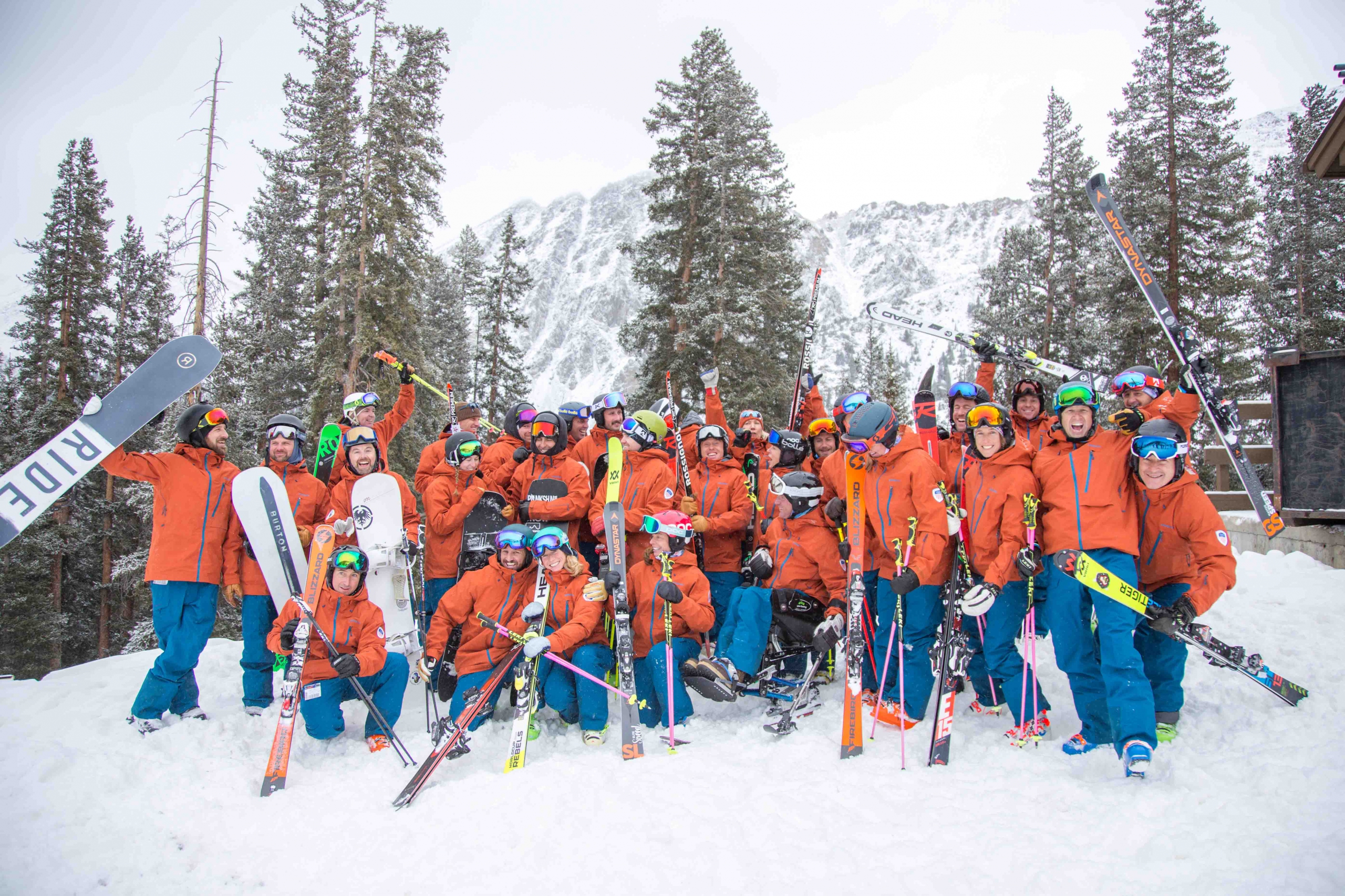 PSIA-AASI National Team celebrates outside in front of snow covered trees holding their skis and snowboards in the air